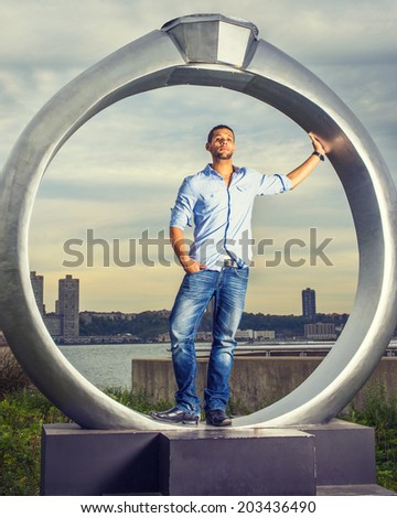 Man Missing You. Wearing a light blue shirt, blue jeans, leather shoes, a young handsome guy with a little beard, mustache is standing by a ring structure, waiting for you, symbolic for engagement.