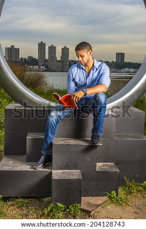 Man Reading Outside. Dressing in a light blue shirt, blue jeans, hands holding a red book, a young college student with a little beard, mustache is sitting by a river, looking down, reading, relaxing.