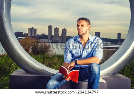 Man Reading Outside. Dressing in light blue shirt, blue jeans, hands holding a red book, a young handsome college student with a little beard, mustache is sitting by river, reading, frowned, thinking.