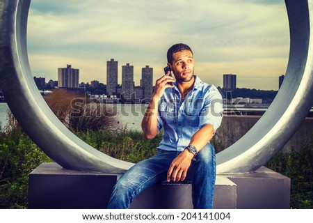Modern Life. Wearing a light blue shirt, blue jeans, a young handsome college student with a little beard, mustache is sitting by river, talking on a mobile phone.