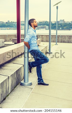 Man Thinking Outside. Wearing a light blue shirt,  blue jeans, leather shoes,  sunglasses hanging on shirt, a young guy with a little beard, mustache is leaning on a pole, sad, frowned, thinking