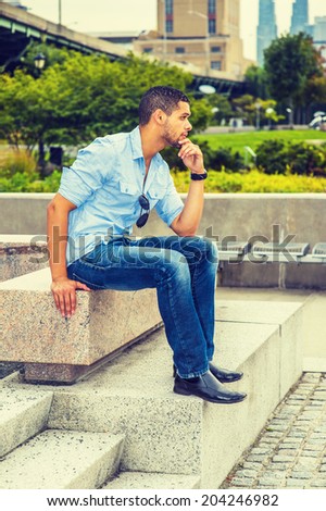 Man Thinking Outside. Wearing shirt, blue jeans, leather shoes, sunglasses hanging on shirt, hand touching his chin, a young guy with a little beard, mustache is sitting on concrete bench, thinking.