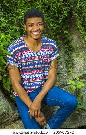 Portrait of Young Black Man. Wearing a short sleeve, collarless, colorful pattern shirt, a young handsome guy is sitting against rocks with green leaves, smiling, charmingly looking at you.