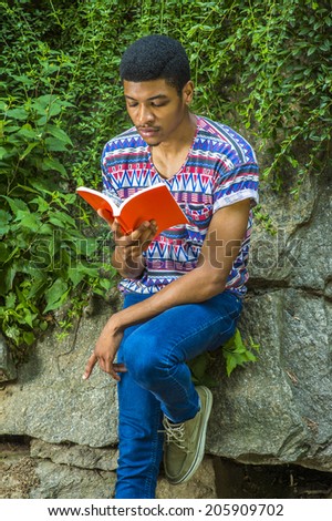 Man Reading Outside. Wearing a short sleeve, collarless, colorful pattern shirt, blue jeans, a young college student is sitting against rocks with green leaves, looking down, reading a red book,