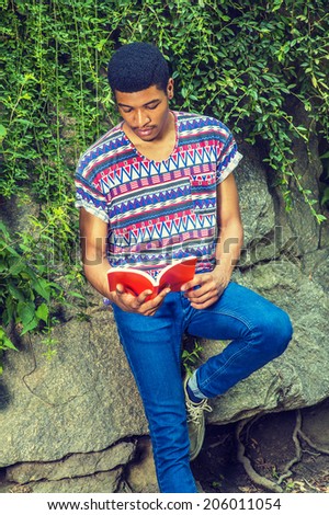 Man Reading Outside. Wearing a short sleeve, collarless, colorful pattern shirt, blue jeans, a young college student is sitting against rocks with green leaves, looking down, reading a red book,