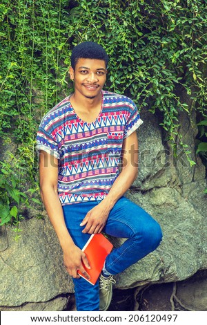 Portrait of Black College Student. Wearing a short sleeve, collarless, colorful pattern shirt, blue jeans, holding a red book, a young guy is sitting against rocks with green leaves, smiling.