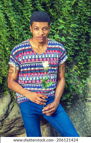Man Waiting for You. Wearing a short sleeve, collarless, colorful pattern shirt, blue jeans, a young black guy is sitting against rocks with green leaves, holding a white rose, looking at you.