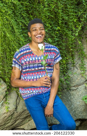Happy Man. Wearing a short sleeve, collarless, colorful pattern shirt, blue jeans, a young black guy is sitting against rocks with green leaves, holding a white rose, opening mouth, laughing.
