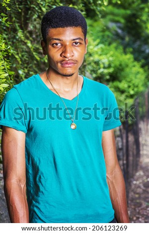 Portrait of Young Black Man. Wearing a green, short sleeve, V-neck T shirt, necklace, a young handsome guy is standing outside in summer, looking at you.