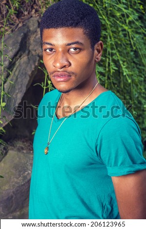 Portrait of Young Black Man. Wearing a green, short sleeve, V-neck T shirt, necklace, a young handsome guy is standing by rocks with green leaves, seriously looking at you.
