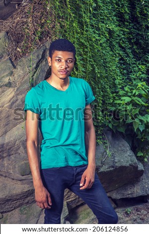 Portrait of Young Black Man. Wearing a green, short sleeve, V-neck T shirt, black pants, a young handsome guy is standing by rocks with green leaves, looking at you.