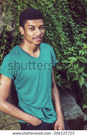 Portrait of Young Black Man. Wearing a green, short sleeve, V-neck T shirt, black pants, a hand in pocket, a young handsome guy is leaning against rocks with green leaves, smiling, looking at you.