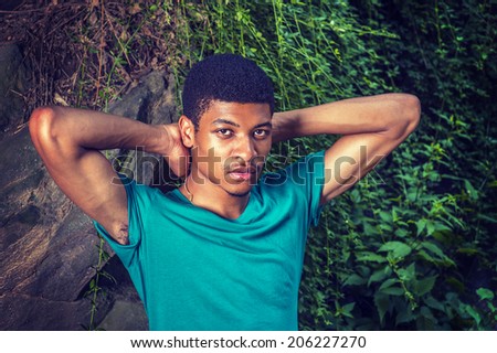 Portrait of Young Black Man. Wearing a green, short sleeve, T shirt,  raising arms, hands in the back of head, a young handsome guy is standing by rocks with green leaves, relaxing.