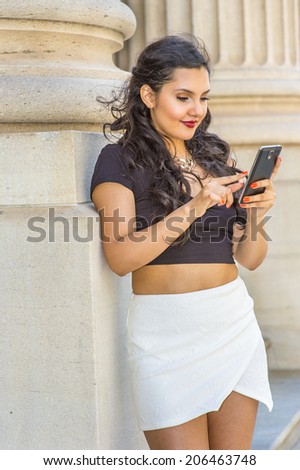 Girl Texting Outside. Dressing in black, short sleeve top, white short wrap skirt, a young lady with long curly hair is checking messages on her mobile phone, fingers touching the screen.