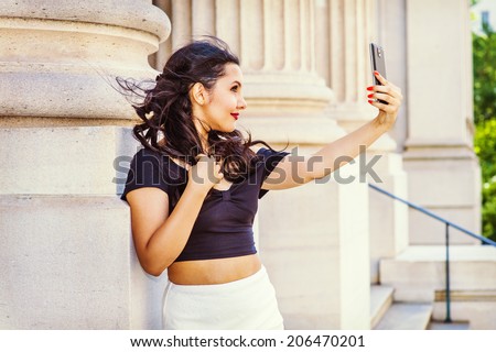 Beautiful girl taken pictures of her self. Stretching arm, holding a mobile device, a young pretty college student is looking through a camera outside an office in the campus. Selfie.