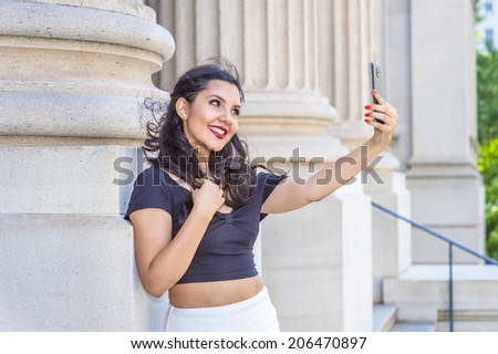 Beautiful girl taken pictures of her self. Stretching arm, holding a mobile device, a young pretty college student is looking through a camera outside an office in the campus. Selfie.