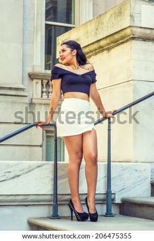College Student. Dressing in black, short sleeve top, white short wrap skirt, high heels, a young pretty lady with long curly hair is standing by railing outside an office, relaxing. Street Fashion
