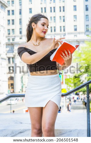 College Student. Dressing in black, short sleeve top, white short wrap skirt, a young pretty girl with long curly hair is standing by railing outside an office, looking down, reading a red book.