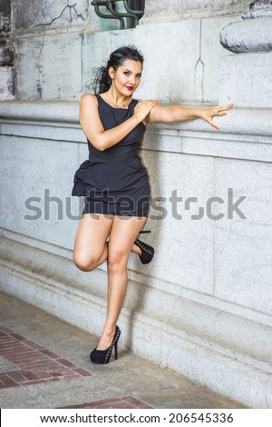 Fashion Girl. Dressing in black, sleeveless top, short pants, high heels, a young pretty lady is standing against an old fashion wall, stretching a arm, bending back a leg, charmingly looking at you.