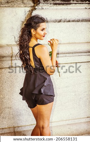 Flower Girl in Back View. Dressing in black, sleeveless top, short pants, a young pretty lady is standing by an old fashion wall, holding a white rose, waiting for you.