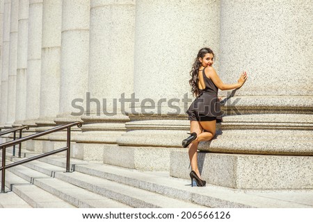 Fashion Girl in Back View. Dressing in black, sleeveless top, short pants, high heels, a young pretty lady with long curly hair is standing by columns,  bending a leg, turning back, looking at you.