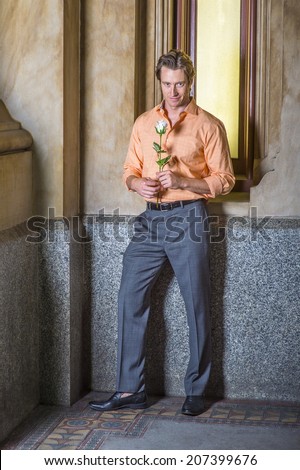 Man waiting for you. Dressing in a light orange patterned shirt, gray pants, leather shoes, hands holding a white rose, a young handsome guy is standing by a small old fashion window, looking at you.