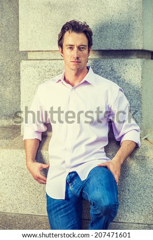 Portrait of Young Man. Wearing a light pink shirt, blue jeans, bending a leg, a young handsome guy is standing against a concrete wall, confidently looking at you.