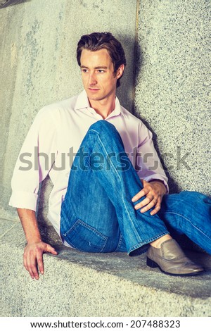 Man Thinking Outside. Wearing a light pink, long sleeve shirt, blue jeans, leather shoes, a young handsome guy is casually sitting against a concrete wall, looking down, thinking