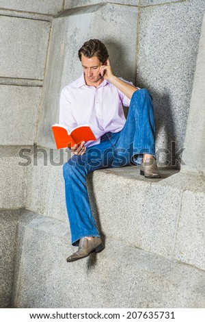 Leisure Time. Wearing a light pink, long sleeve shirt, blue jeans, leather shoes, a young guy is casually sitting against a concrete wall, reading a book, making a phone call at the same time.