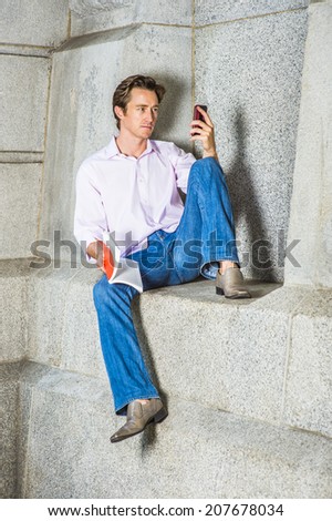 Relaxing Outside. Wearing a light pink, long sleeve shirt, blue jeans, leather shoes, a young man is casually sitting against a concrete wall, holding a red book, checking messages on his smart phone.