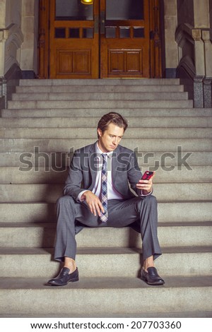 Businessman Waiting for You. Dressing formally in gray suit, white under shirt, patterned neck tie, a young businessman is casually sitting on steps outside an office, texting on his mobile phone.