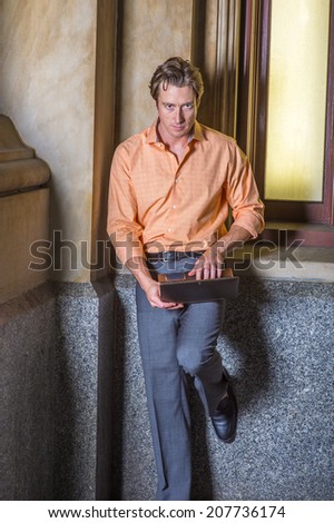 Man Working Outside. Dressing in a light orange patterned shirt, gray pants, a young handsome guy is standing by an old fashion window in the corner, working on a laptop computer.