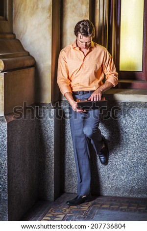 Man Working Outside. Dressing in a light orange patterned shirt, gray pants, leather shoes, a young guy is standing by an old fashion window in the corner, looking down, working on a laptop computer.