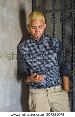 Unhappy Man. Wearing a black patterned shirt, yellow pants, a young guy with beard, yellow hair is leaning against the wall outside a metal gate, looking down on his fingers, sad, thinking.