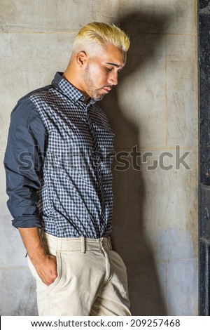 Wearing a black patterned shirt, yellow pants, a young man with beard, yellow hair is leaning against the wall outside a metal gate, hands in pockets, looking down, sad, thinking.