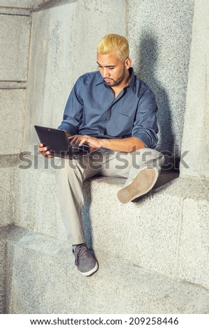 Young Man Working Outside. Wearing a blue shirt, gray pants, casual shoes, a young guy with beard, yellow hair is sitting by a concrete wall, looking down, working on a laptop computer.
