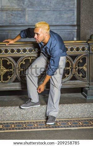 Man Looking for You. Wearing a blue shirt, gray pants, casual shoes, a young guy with beard, yellow hair is standing by old fashion style railing in a hallway, bending back forward, looking around.