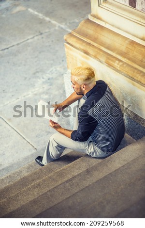 Young Man Reading Outside. Wearing a black patterned shirt, grey pants, leather shoes, a young guy with beard, yellow hair, is sitting on stairs, reading a book.