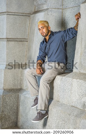 Young Man Relaxing Outside. Wearing a blue shirt, gray pants, casual shoes, a young guy with beard, yellow hair is sitting on a concrete wall, looking at you.