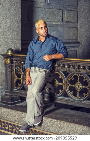 Man Waiting for You. Wearing a blue shirt, gray pants, casual shoes, a young guy with beard, yellow hair is standing by old fashion style railing in a hallway, looking around, relaxing.