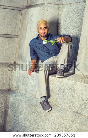 Young Man Waiting Outside. Wearing a blue shirt, gray pants, casual shoes, a young guy with beard, yellow hair is sitting down on a concrete wall, holding a white rose, relaxing, thinking.