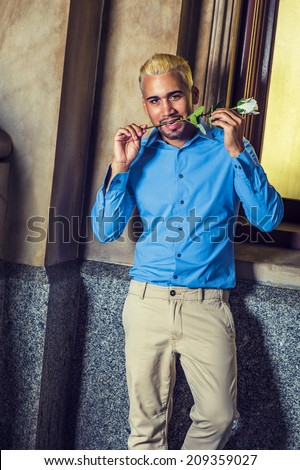 Young Man Waiting for You. Wearing a blue shirt, grey pants, a young guy with beard, yellow hair, is standing by old fashion window, a white rose keeping in mouth, smiling, looking at you.