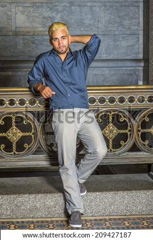 Man Waiting for You. Wearing a blue shirt, gray pants, casual shoes, a young guy with beard, yellow hair is standing by old fashion style railing in a hallway, looking at you.