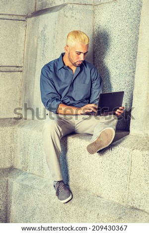 Young Man Working Outside. Wearing a blue shirt, gray pants, casual shoes, a young guy with beard, yellow hair is sitting by a concrete wall, looking down, working on a laptop computer.