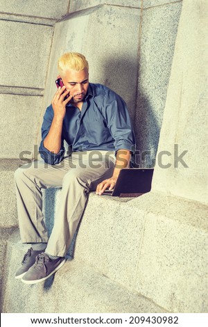 Man Working Outside. Wearing a blue shirt, gray pants, casual shoes, a young guy is sitting by a concrete wall, looking down, working on a laptop computer, talking on a mobile phone in the same time.