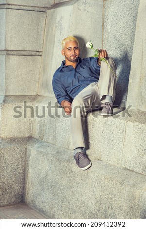 Young Man Waiting Outside. Wearing a blue shirt, gray pants, casual shoes, a young guy with beard, yellow hair is sitting down on a concrete wall, holding a white rose, relaxing, thinking.