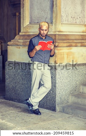 Young Man Reading Outside. Wearing a black patterned shirt, grey pants, leather shoes, hands holding a red book, a young guy with beard, yellow hair, is standing against a column, reading.