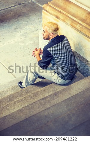 Young Man Relaxing Outside. Wearing a black patterned shirt, grey pants, leather shoes, a young guy with beard, yellow hair, is sitting on stairs, back view.