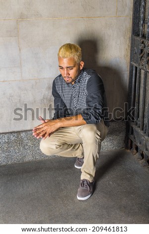 Unhappy Man Thinking on the Corner. Wearing a black patterned shirt, yellow pants, casual shoes, a young guy is squatting against the wall outside a metal gate, looking down, sad, thinking.