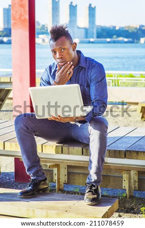Man Working on Site. Wearing a blue shirt, pants, leather shoes, a hand touching his chin, a young black guy with mohawk hair is sitting on a wooden structure by a river, working on a laptop computer.